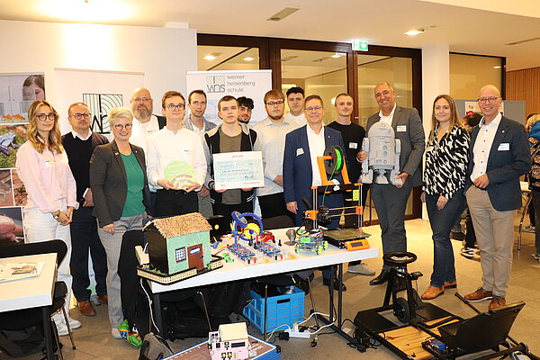 Gruppenfoto nach der Auszeichnung Gruppenfoto bei der 21. MINT-Messe: Mehrere Personen stehen hinter einem Tisch mit technischen Exponaten wie 3D-Druckern, Robotermodellen und einem Miniaturhaus. In der Mitte hält eine Person einen großen symbolischen Scheck, daneben eine runde Auszeichnung mit der Aufschrift ‚Provadis MINT 2025‘. Im Hintergrund sind Banner der Werner-Heisenberg-Schule zu sehen