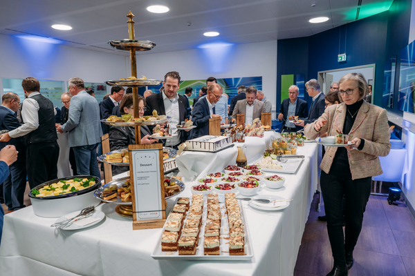  Tisch mit Dessertbuffet und Snacks, umgeben von Personen in Business-Kleidung, die sich unterhalten und Speisen nehmen.