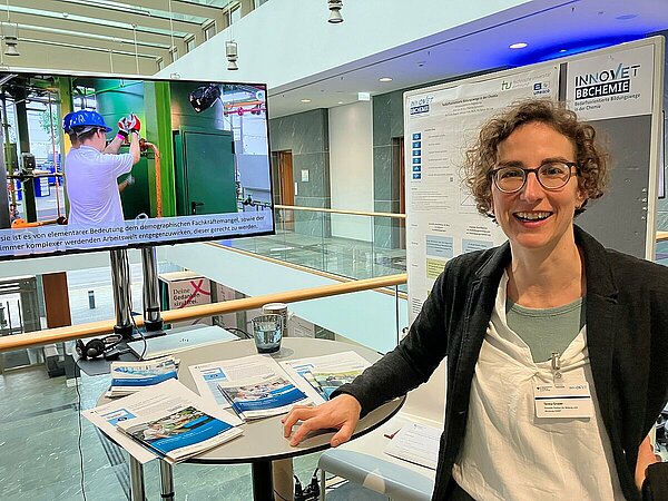 Frau lächelt an Berufsbildungsstand auf Messe. Frau an einem Informtionsstand mit Poster.