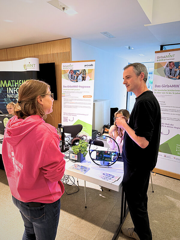 Austausch am Girls4MINT-Stand Zwei Personen stehen an einem Informationsstand mit Bannern zum Girls4MINT-Programm. Auf dem Tisch liegen Flyer, eine Pflanze und ein technisches Gerät mit Kabeln und Platine. Im Hintergrund sind Roll-Ups mit Texten zu MINT-Angeboten sichtbar.